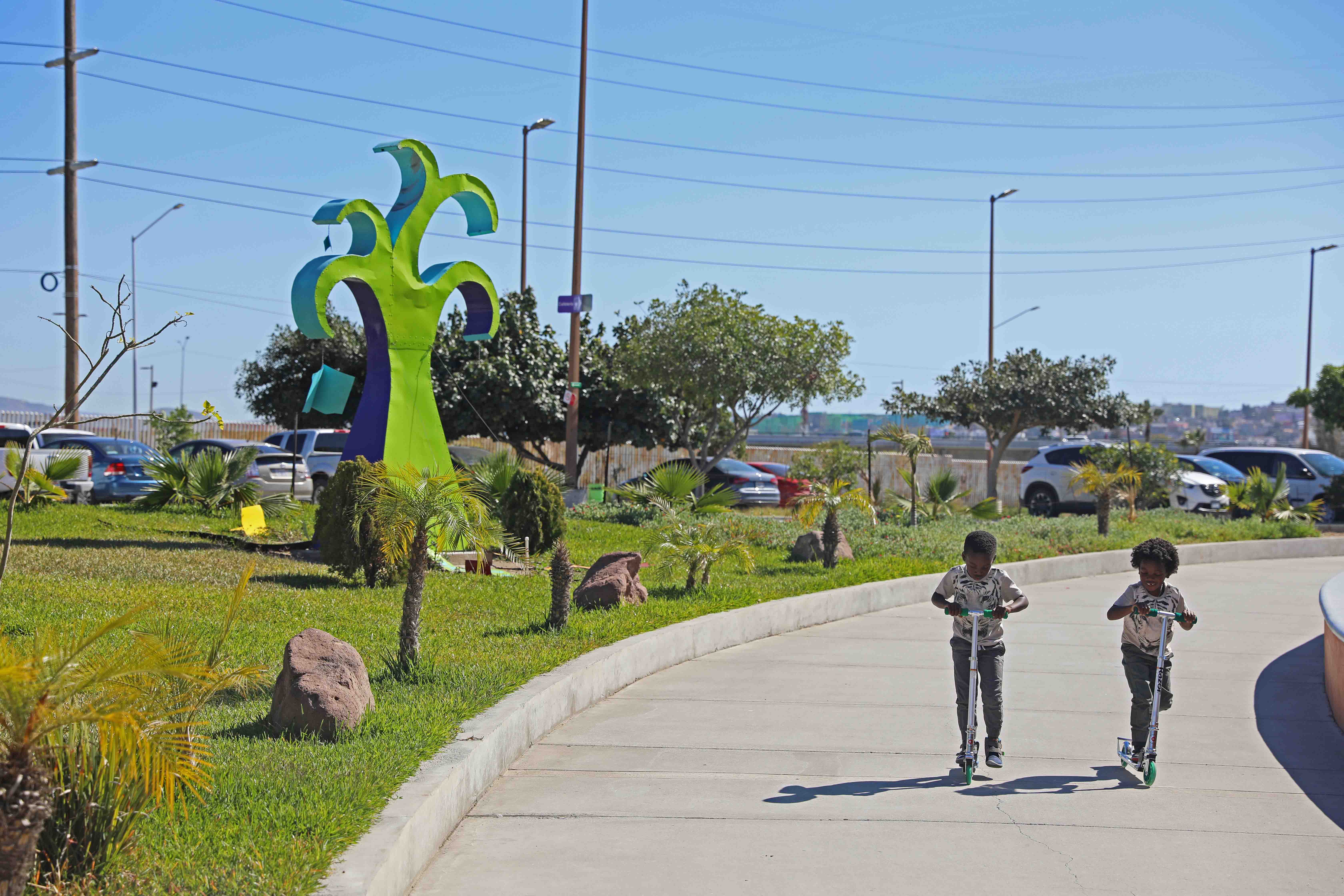 Two boys riding scooters past a piece of public art in Tijuana