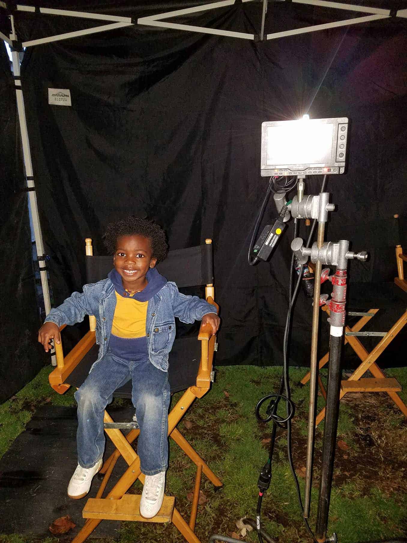 Child model smiling at the camera. while sitting in a director chair with a black background