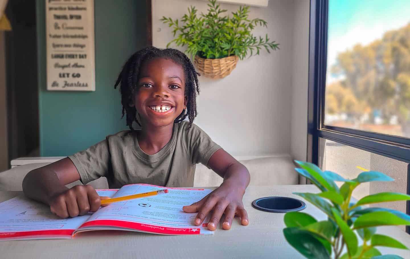 A young boy sits at a desk with a school workbook open smiling and holding a pencil enjoying his homeschooling lesson.